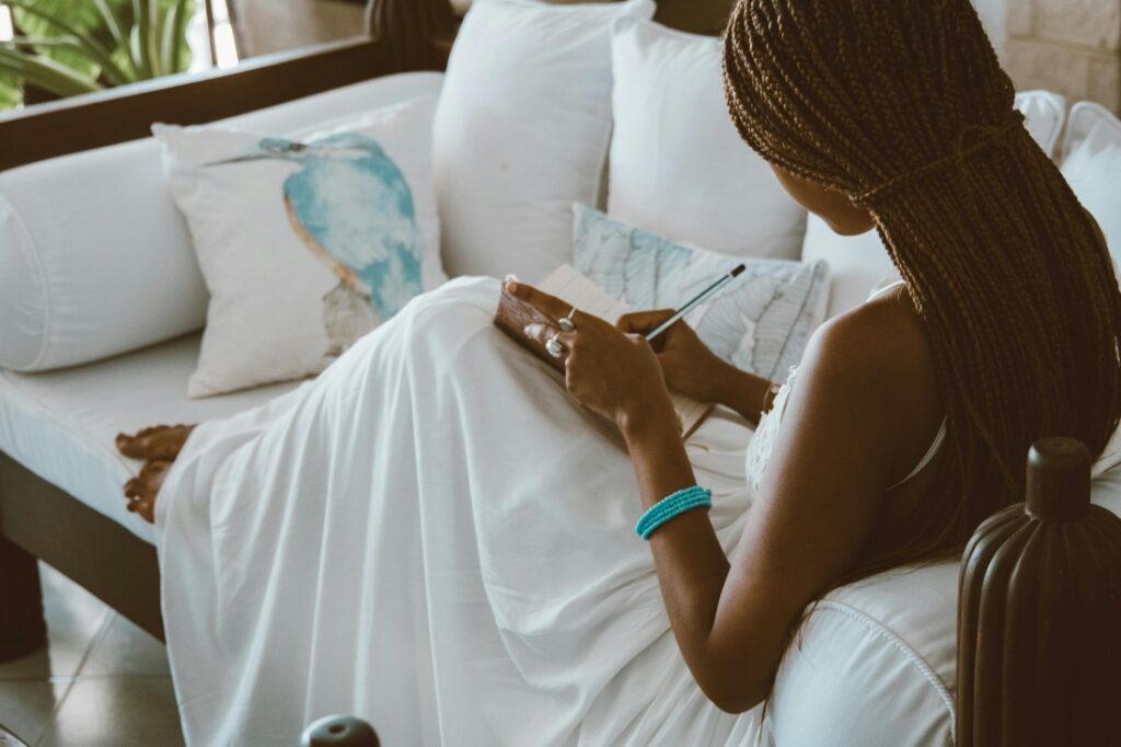 A woman with braids, wearing a white dress and a turquoise beaded bracelet, sitting on a white cushioned sofa while journaling in a notebook.