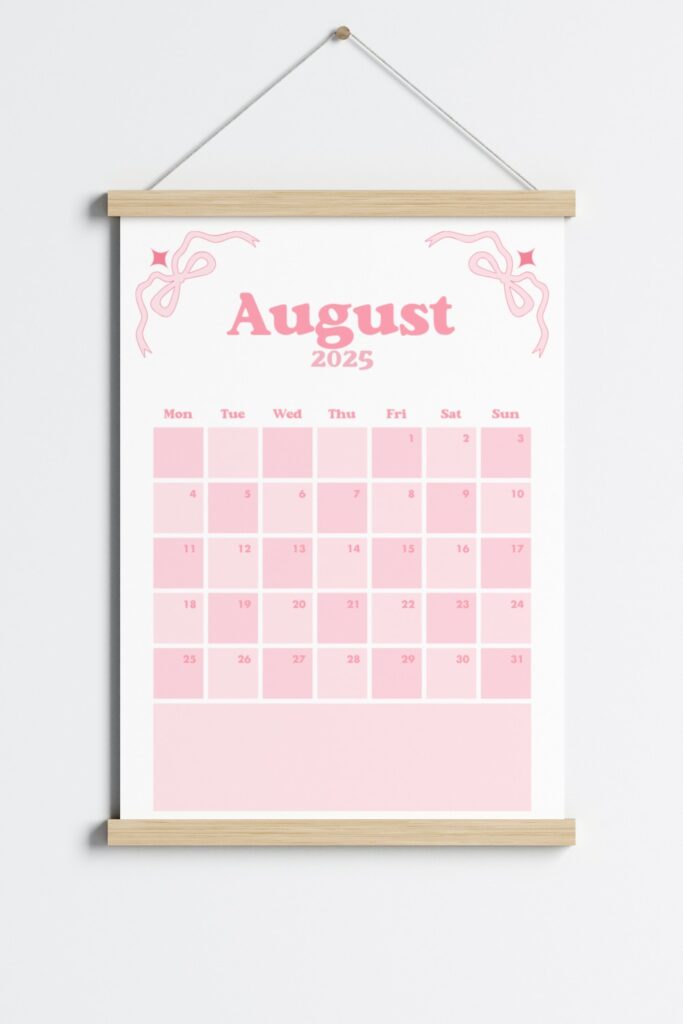August 2025 calendar page in a pink coquette theme, featuring bows in the corners, a Monday start layout and displayed in a wooden hanging frame on a light grey wall.