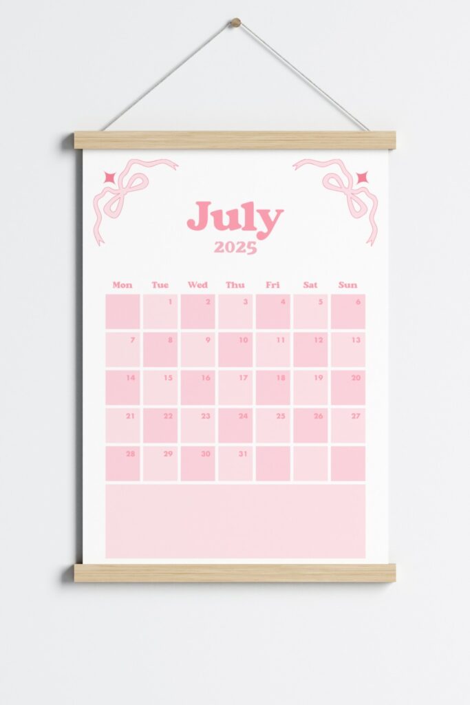 July 2025 calendar page in a pink coquette theme, featuring bows in the corners, a Monday start layout and displayed in a wooden hanging frame on a light grey wall.