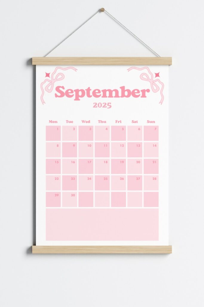 September 2025 calendar page in a pink coquette theme, featuring bows in the corners, a Monday start layout and displayed in a wooden hanging frame on a light grey wall.