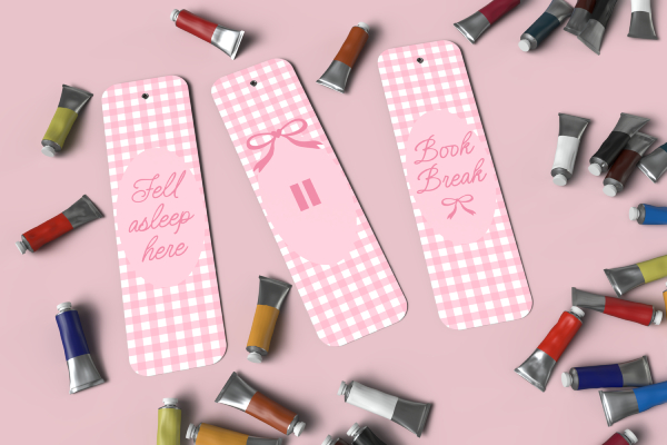 A mockup of three pink coquette-themed bookmarks with a gingham pattern. The bookmarks say "Fell asleep here" and "Book Break" and are scattered on a pink surface with paint tubes.