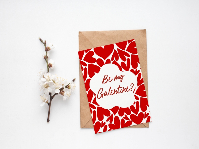 A Galentine's card with a bold red heart pattern and a white center cloud shape that reads "Be my Galentine?" in red script. THe card sits on top of a brown envelope on a white surface with white flowers beside it