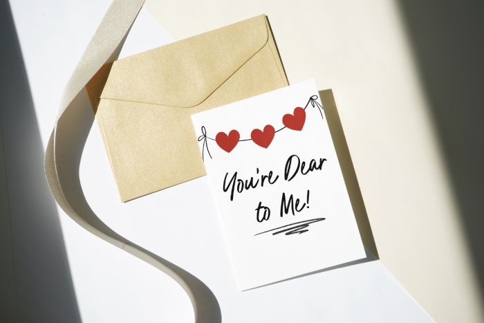 A white Galentine's card with red heart bunting that says "You're Dear to Me!" placed next to a gold-brown kraft envelope and a flowing satin ribbon.